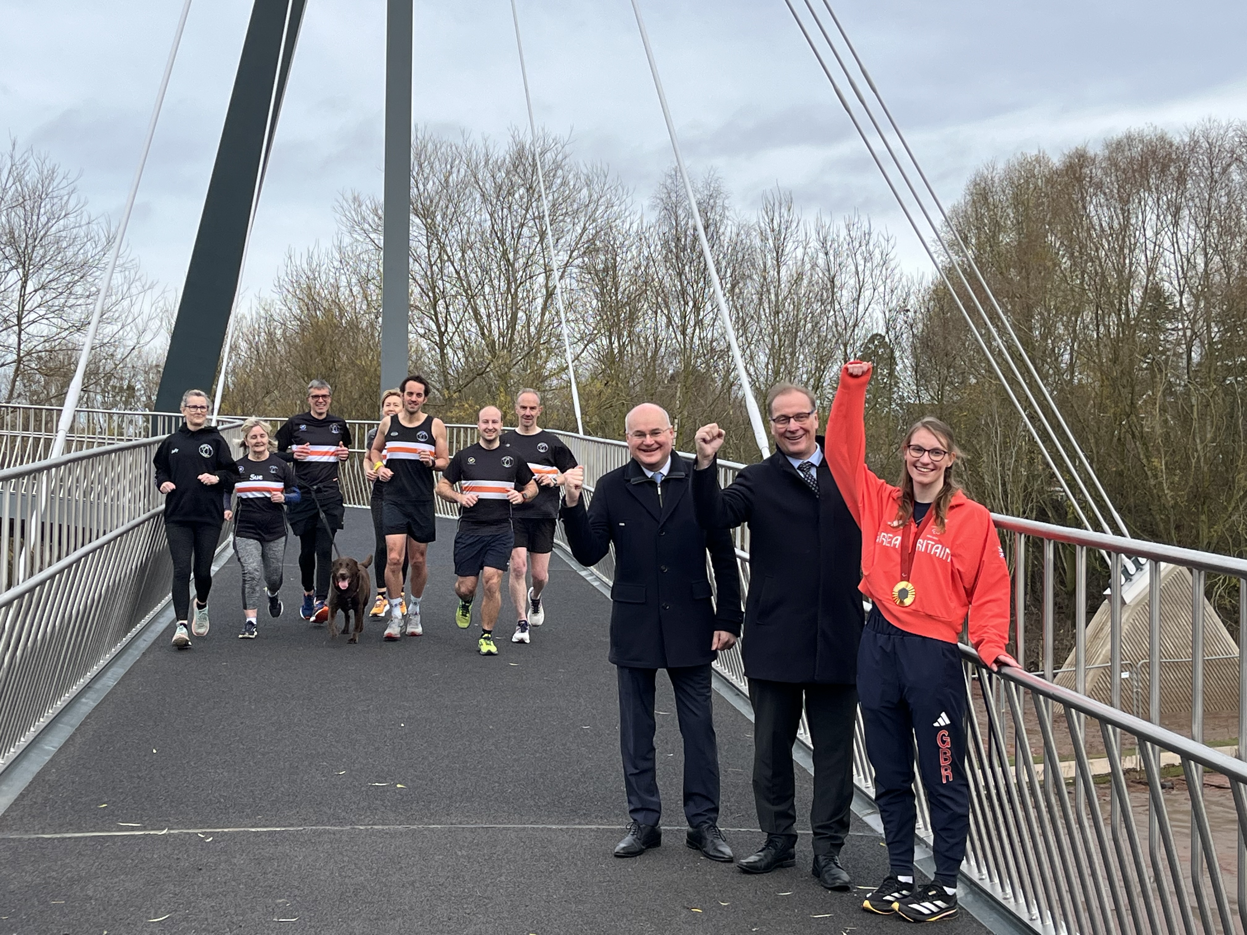 Councillor Simon Geraghty, Councillor Marc Bayliss and Rebecca Redfern MBE stood on the bridge with members of the Black Pear Joggers running towards the camera in the distance.