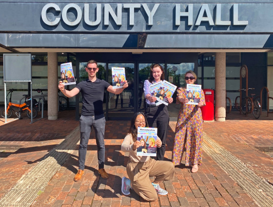 4 people stand outside the entrance to County Hall, each holding a copy of the INclusive Worcestershire magazine. There are 3 people stood at the rear (a man to the left and 2 women to his right) and a young woman kneels in the foreground facing the camera. 