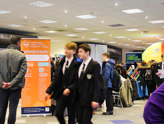 School students attend a carers event at Worcester's Sixways Stadium. they are walking across an exhibition hall with exhibitors in the background.